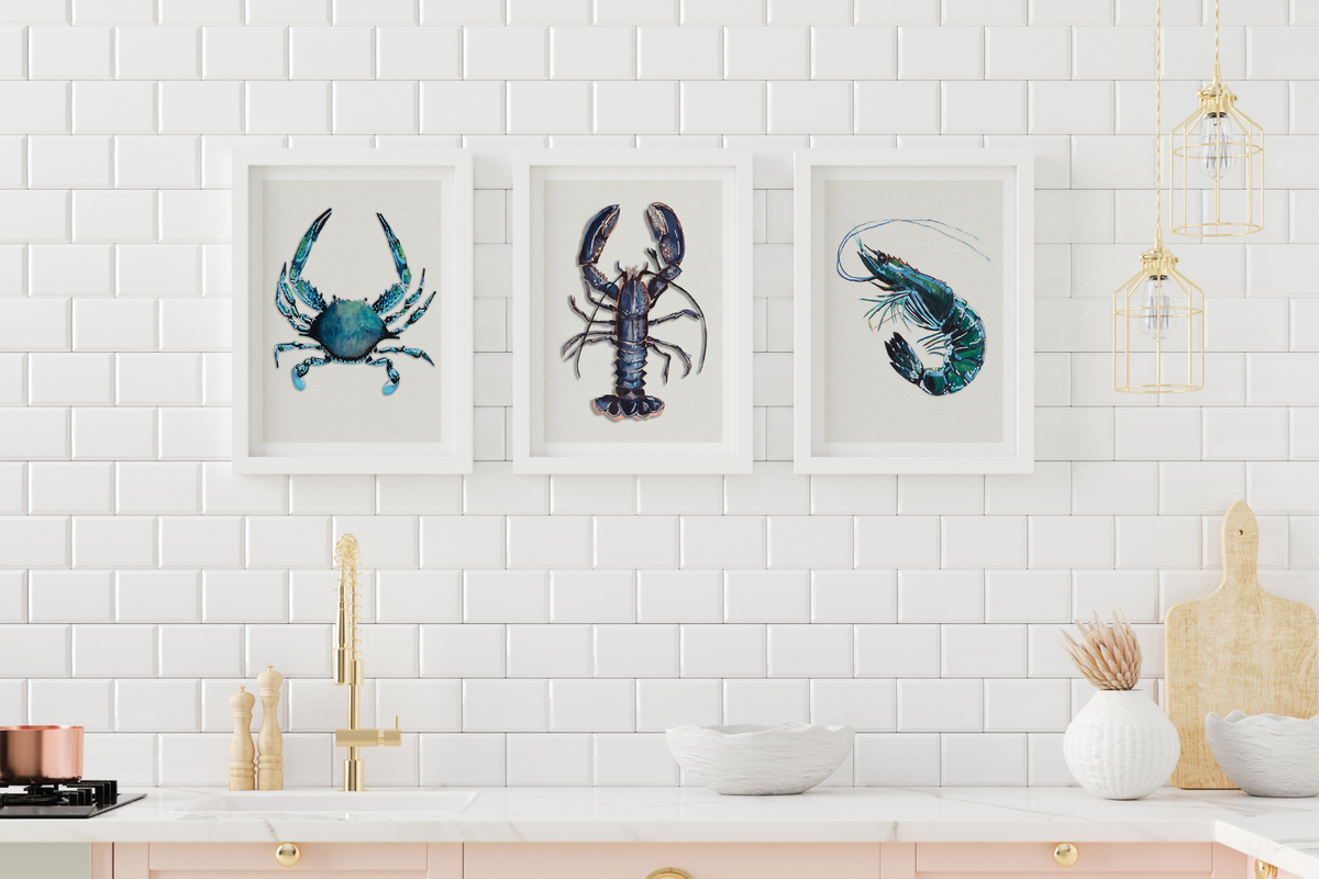 Set of three shellfish kitchen art prints, featuring a blue crab, a lobster painting, and a prawn, elegantly framed and displayed on a kitchen wall above a wooden countertop, adding a marine-inspired touch to culinary spaces.