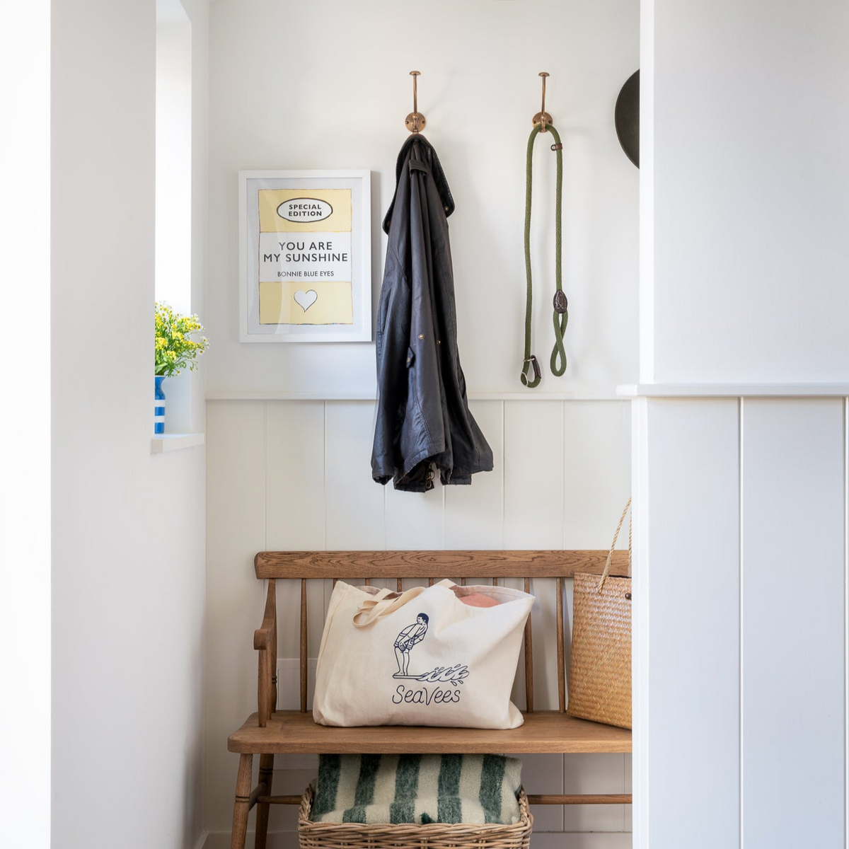 Yellow quote art print in hallway above bench. Shows a art print in bootroom by brass coat hooks aove wooden bend. Bootroom has half height panelling in off white.