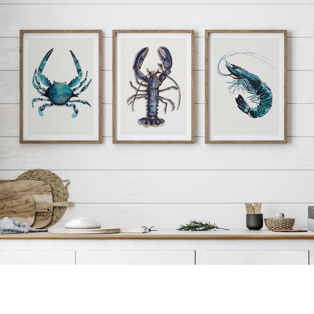 Set of three shellfish kitchen art prints, featuring a blue crab, a lobster painting, and a prawn, elegantly framed and displayed on a kitchen wall above a wooden countertop, adding a marine-inspired touch to culinary spaces.