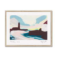 A framed abstract landscape art print featuring a stylized coastal scene with blue mountains, pink and white foreground shapes, dark brown rock forms, and a pale yellow band near the horizon. The print is mounted with a wide white mat and enclosed in a light natural wood frame, shown against a plain white background.