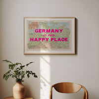 Framed map with 'Germany is our Happy Place' text on a wall with a plant and chair below.