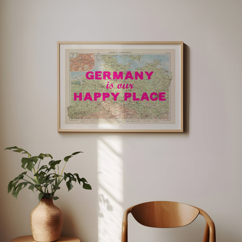 Framed map with 'Germany is our Happy Place' text on a wall with a plant and chair below.