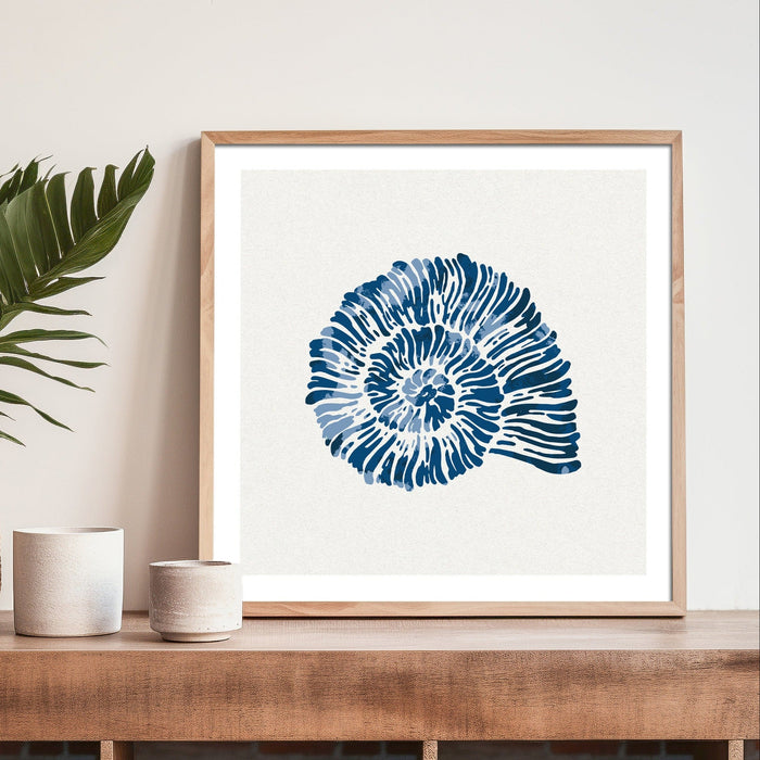 Framed shell art print - indigo spiral shell wall art in coastal interior, styled on wooden console with plant and ceramics.