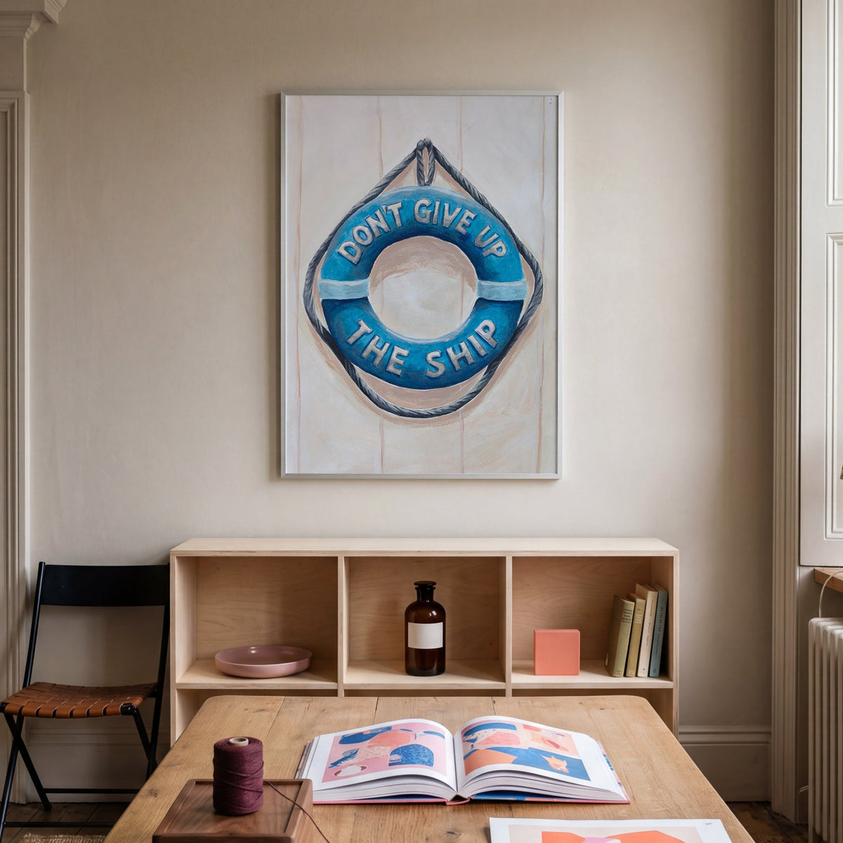 Nautical painting with a life buoy and a motivational quote in a room interior with a wooden table, a shelf, and wall art.