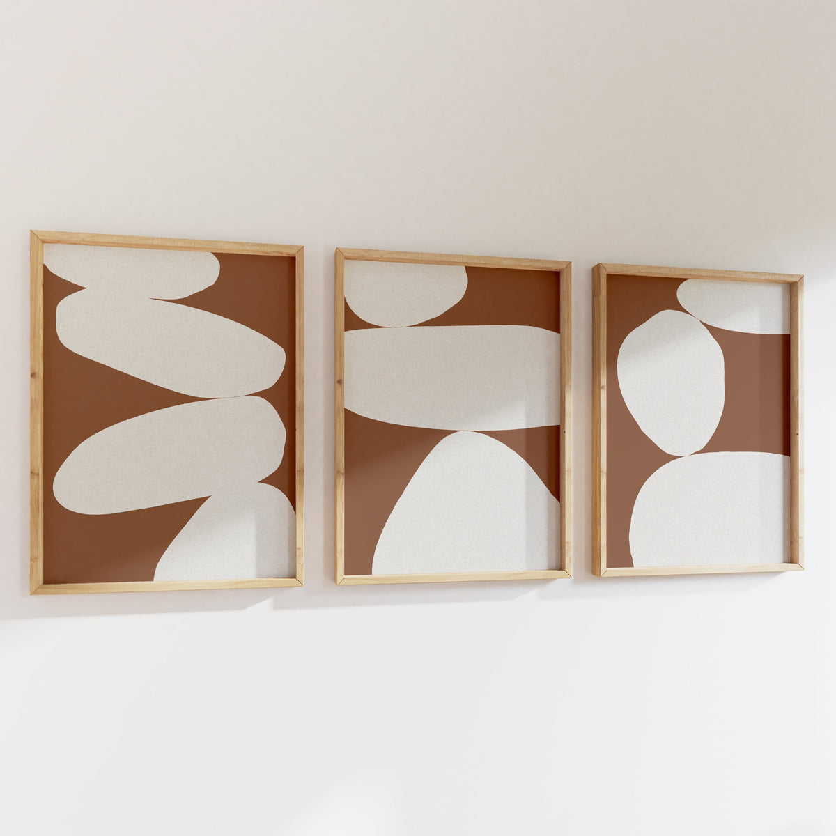 Three abstract wall art pieces with white shapes on a brown background, framed in wooden frames, mounted on a white wall.
