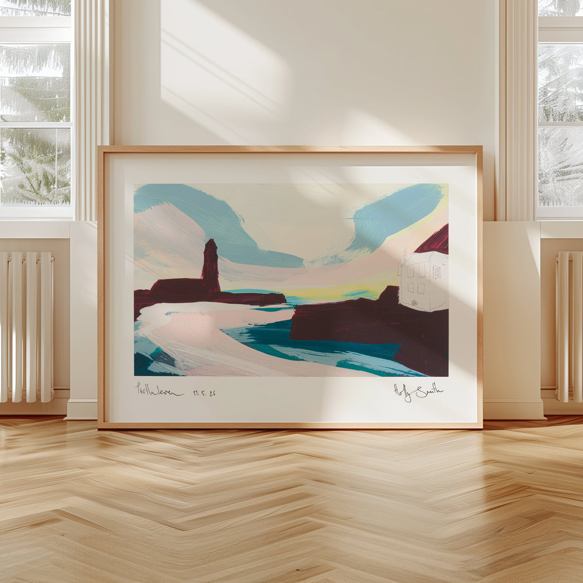 A framed abstract landscape art print leaning against a wall in a bright room with wood floors and large windows.