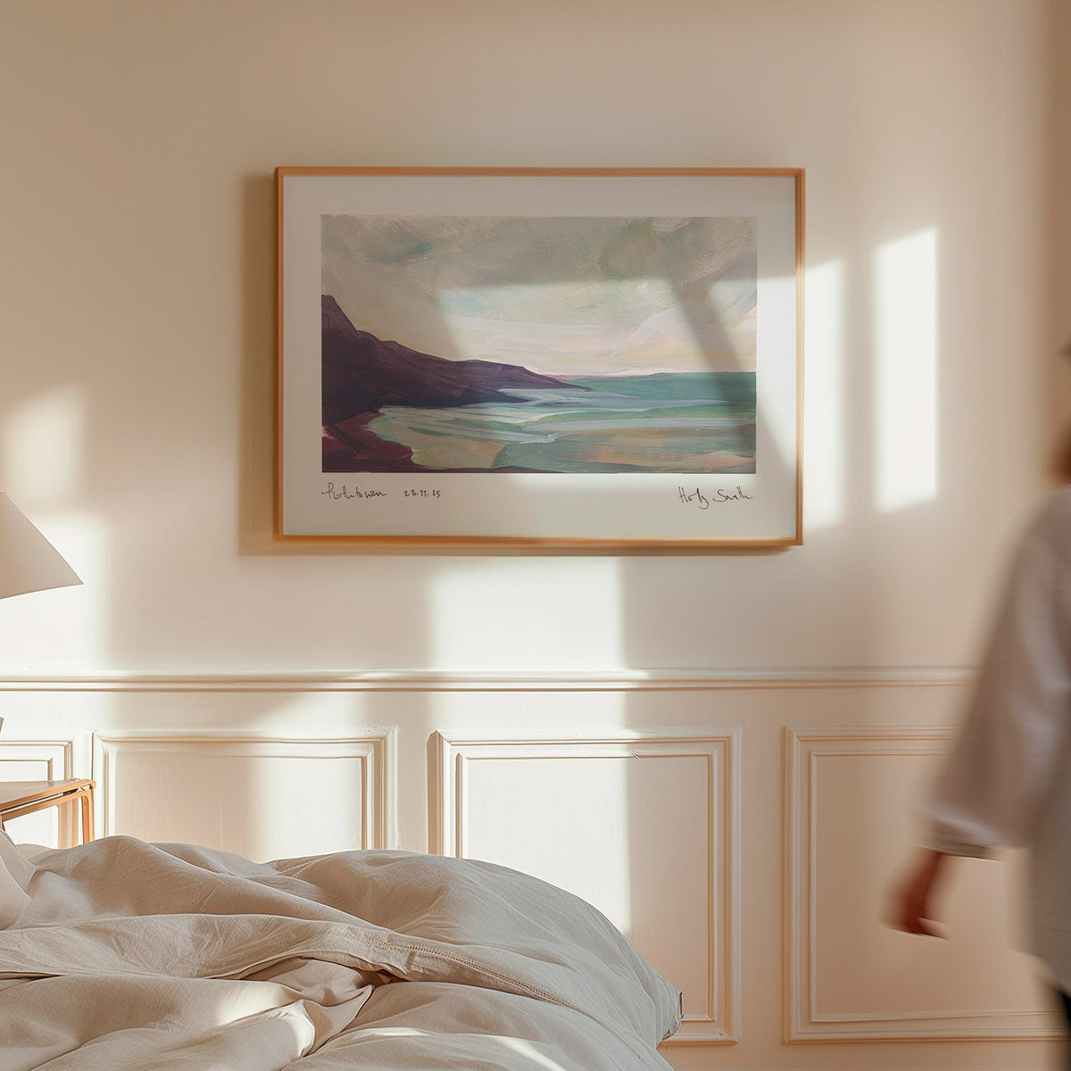 A framed landscape art print of Porthtowan in Cornwall in a modern bedroom. Featuring an impressionistic painting of a coastal scene with dark cliffs, turquoise water, and a cloudy sky, shown against a plain white background.
