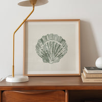 Framed artwork of a shell on a wooden desk with a lamp and books.