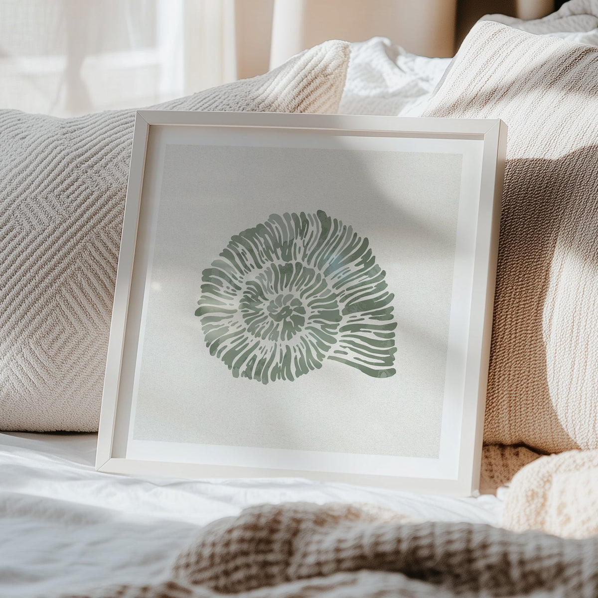 Shell art print in a white wood frame styled in a bright coastal interior. Framed shell artwork with a green design on a bed with beige pillows and blanket.