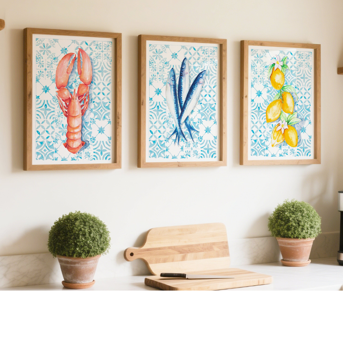 Three framed artworks of a lobster, blue shellfish, and lemons on a wall above a kitchen counter with plants and cutting boards.