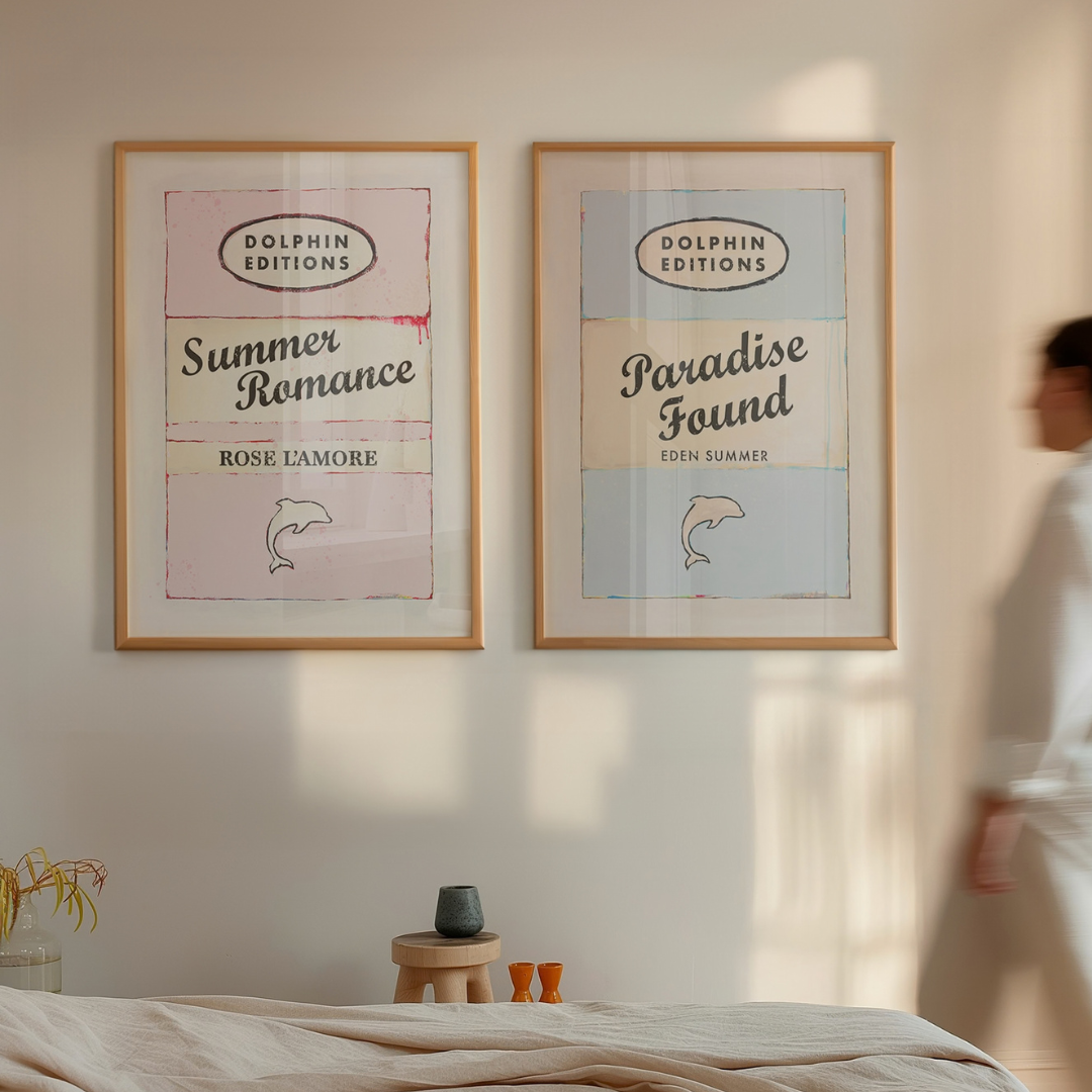 Two framed books prints 'Summer Romance' and 'Paradise Found' on a wall with a person walking by.
