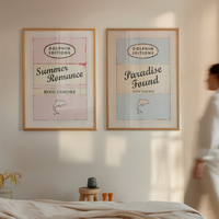 Two framed books prints 'Summer Romance' and 'Paradise Found' on a wall with a person walking by.