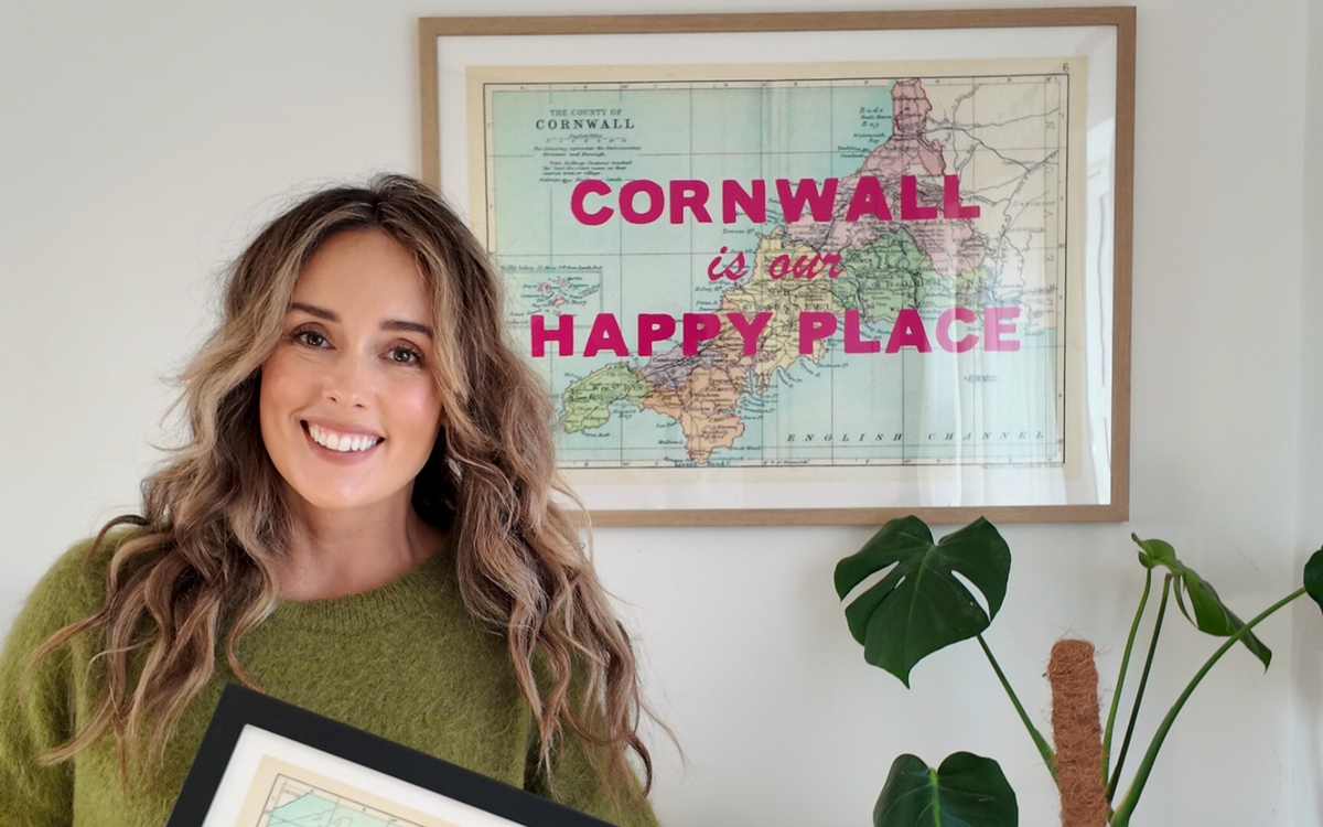 Woman holding a framed map print with 'Cornwall is our happy place' text. Personalised map print and map art