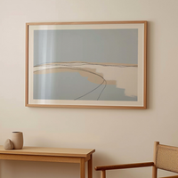 Framed abstract coastal artwork on a wall with a wooden table and chair in the foreground.