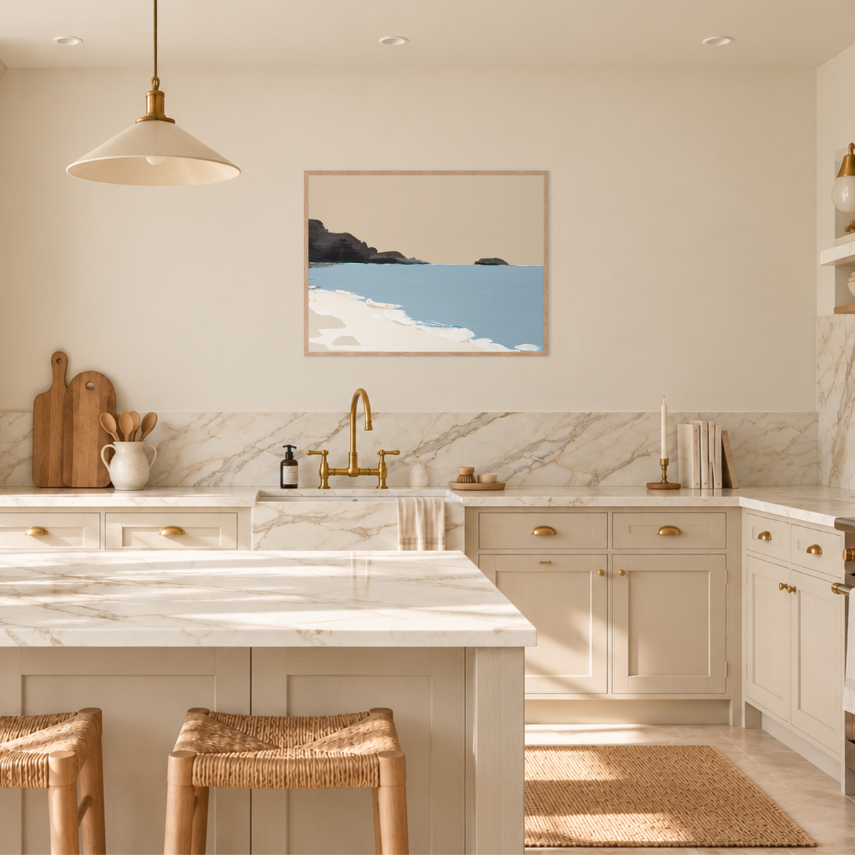 A modern kitchen interior with cream cabinetry, white marble countertops and backsplash, brass fixtures, pendant lights, woven bar stools, and a framed coastal-style wall art print above the sink.