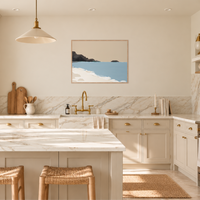 A modern kitchen interior with cream cabinetry, white marble countertops and backsplash, brass fixtures, pendant lights, woven bar stools, and a framed coastal-style wall art print above the sink.