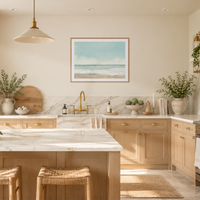 A bright, modern kitchen interior with light wood cabinetry, a white marble island, a brass faucet, and a framed ocean painting centered above the sink.