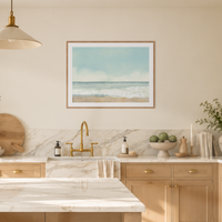 A framed art print depicting a calm ocean beach scene hangs on a beige wall above a marble sink and light wood kitchen cabinets.
