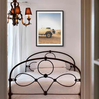 a photograph of land rover defenders on the beach ealry in the morning, in a black frame above a victorian bed in a small bedroom with a victorian chandelier
