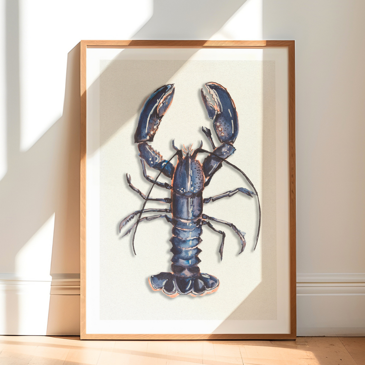 Blue lobster art print in a natural oak frame, styled as coastal kitchen wall art in a bright, sunlit room.