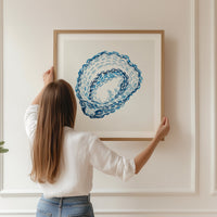 Oyster shell coastal wall art featuring layered blue brushstrokes on a soft neutral background. Framed and being hung in living room