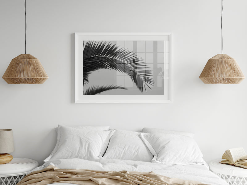 a framed print of a palm leaf above a bed. This peice of bedroom art is in a white painted bedroom and the bed is mae with white linen sheets and white bedside tables