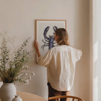 lady hanging a lobster art print on wall in dining room. lobster painting framed with natural wood frame.