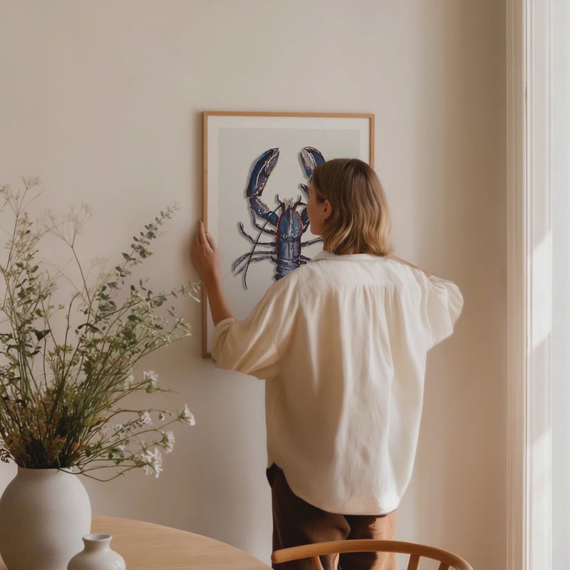 lady hanging a lobster art print on wall in dining room. lobster painting framed with natural wood frame.