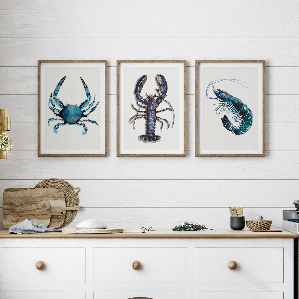 set of three shellfish prints in a white kitchen above a worksurface. Lobster print, crab painting and prawn painting.