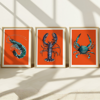 Set of three shellfish art prints - colourful kitchen at of three framed artworks of a prawn, lobster, and crab on orange backgrounds.