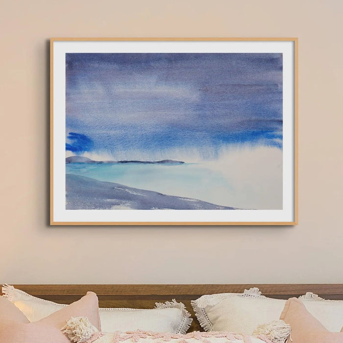 blue seascape painting framed, with headland and beach in foreground. the watercolour beach paitning is framed in a natural wood frame and is above a bed